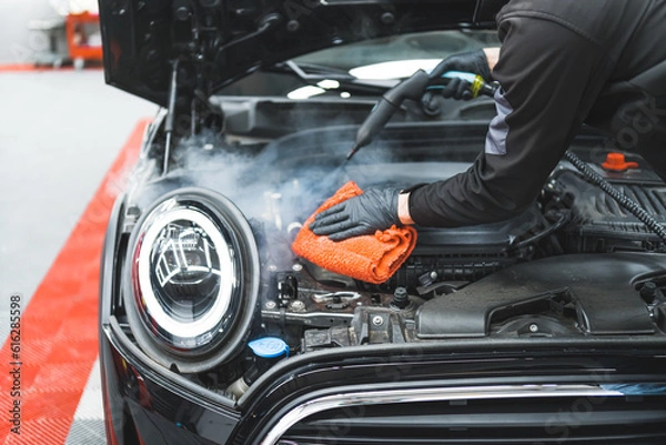Fototapeta Car detailing concept. Manual engine cleaning with steam. Car detailing expert deep-cleaning black vehicle and wiping car surface with orange microfibre cloth. High quality photo