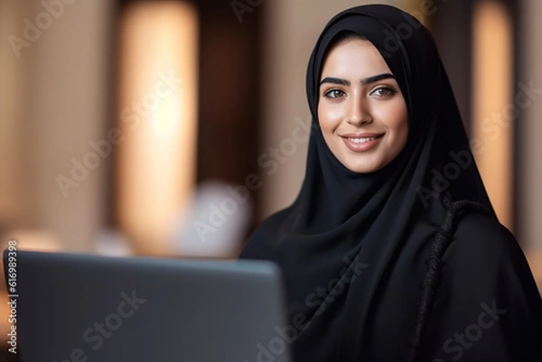 Fototapeta Portrait of beautiful smiling Arabic woman in Abaya working on laptop and looking to camera at office.
