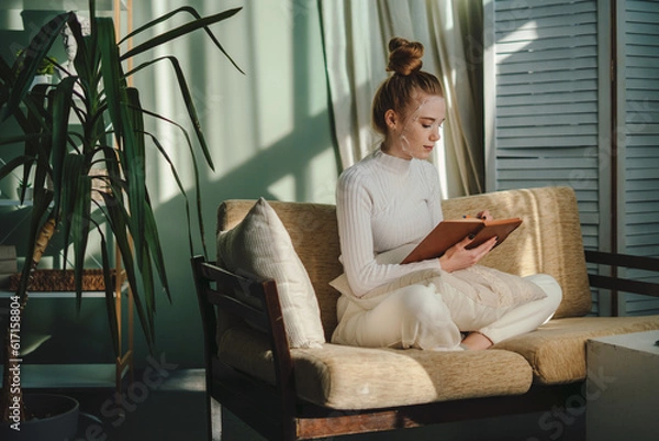 Fototapeta Side view of young female freelancer in casual clothes sitting on sofa and reading notes in notebook while working on remote project