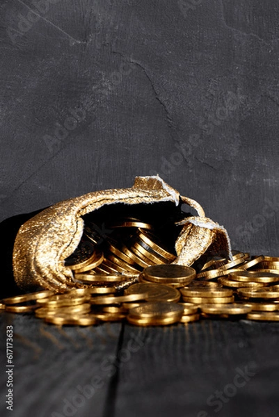 Fototapeta A bag with gold coins on a wooden background. The money is in a black bag. Treasure Hunt. A Purse with money on a black background. Wallet wit gold coins.Copy space