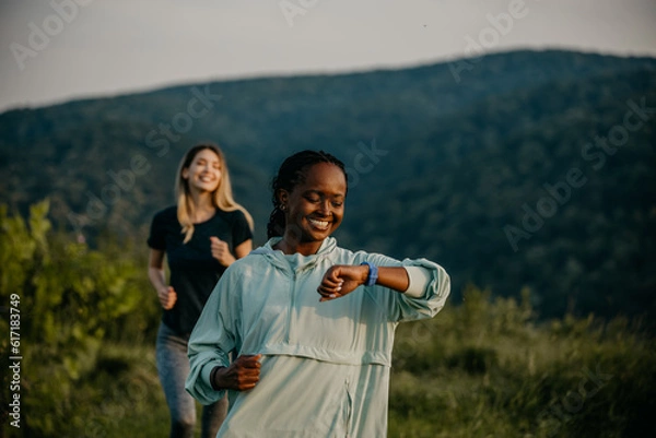 Obraz Active Lifestyle in the Wilderness: Embracing an active lifestyle, black and white female joggers navigates a trail in a lush forest, using their smartwatch to maintain their pace and monitor their ov