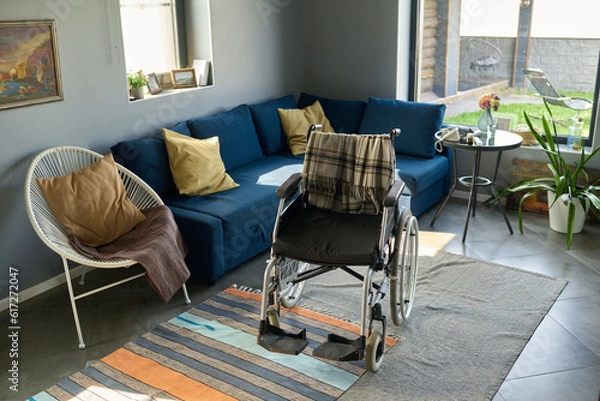 Fototapeta Wheelchair with woolen plaid standing on handmade rug in the center of living room surrounded by comfortable couch and armchair with cushions