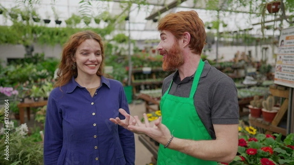 Fototapeta A male florist speaking with female customer inside Flower Shop. Local Business concept of worker wearing green apron assisting client