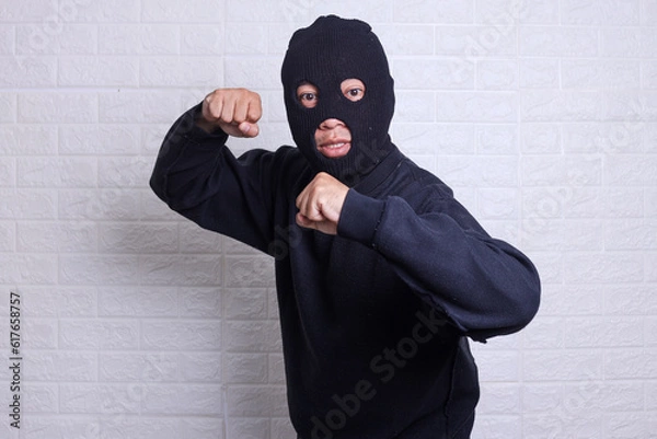 Fototapeta Angry criminal menacing fist to the camera ready to fight isolated on white background. Delinquent wearing face mask.