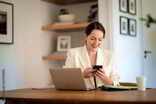 Fototapeta Attractive young woman using mobile phone and laptop and working at home