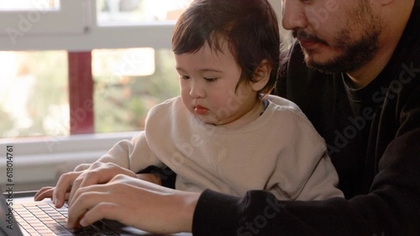 Obraz A young man trying to work on laptop holding his son at home. Remote work