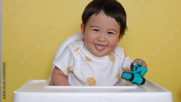 Obraz Cute asian baby sits on highchair and smiling on yellow background