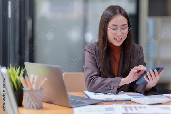 Fototapeta Sharing good business news. Attractive young businesswoman talking on the mobile phone and smiling while sitting at her working place in office and looking at laptop PC.