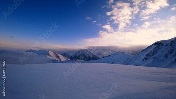 Obraz An ocean of clouds in the snowy mountains at dawn. White clouds are like a carpet in a gorge. Waves rise and descend from the mountains. Sunrise of the yellow sun. Clear sky, mountain peaks. Almaty