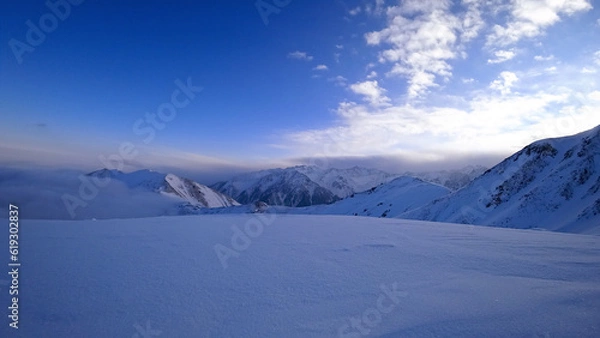 Obraz An ocean of clouds in the snowy mountains at dawn. White clouds are like a carpet in a gorge. Waves rise and descend from the mountains. Sunrise of the yellow sun. Clear sky, mountain peaks. Almaty