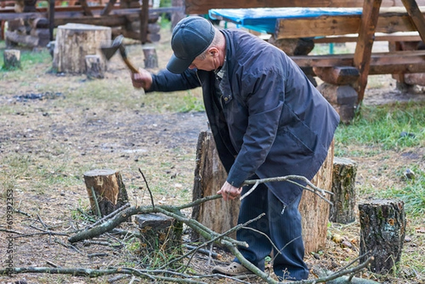Obraz Elderly retired man chopping firewood with an ax, cooking grilled food on a fire