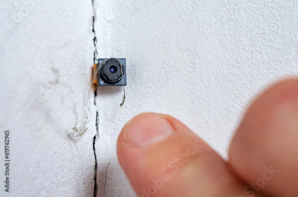 Fototapeta Spy hidden camera in the crack of the wall. Man discovers micro camera in office. Spy scandal, collecting compromising evidence, wiretapping, symbolic image.