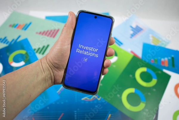 Obraz Ręka trzymająca telefon na którym jest wyświetlany napis investor relations na tle plansz z wykresami.
