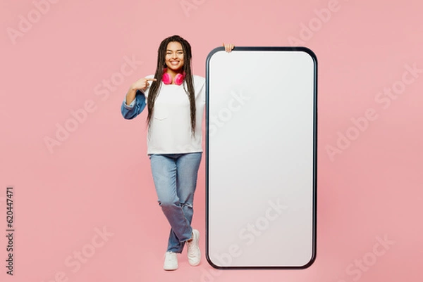 Obraz Full body young woman of African American ethnicity wear white sweatshirt casual clothes point finger on big huge blank screen mobile cell phone smartphone with area isolated on plain pink background.