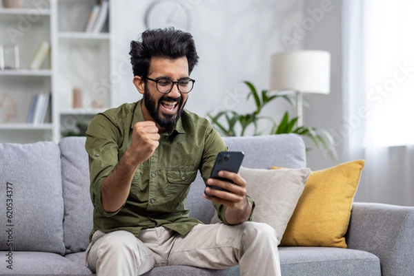 Fototapeta Happy and satisfied with achievement results hispanic at home reading online win notification in online application on phone, man smiling holding hand up triumph gesture sitting sofa in living room.