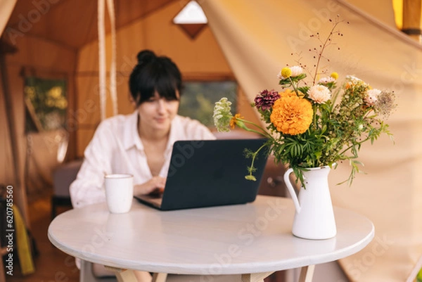 Fototapeta female traveler sitting beside camping tent, drinking morninf coffee and using notebook laptop computer working