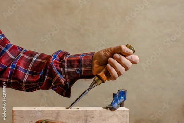 Fototapeta Close-up of the hand of a skilled craftsman holding a chisel as he firmly drives it into a piece of wood