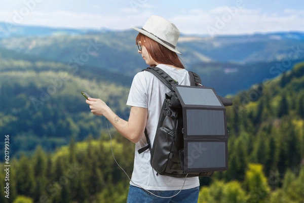 Fototapeta Young woman with mobile phone in hands connected to backpack with portable solar battery