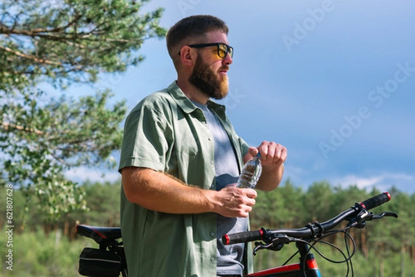 Fototapeta Cyclist have rest near the bicycle, in the forest and relax for energy in adventure workout, drinking bottle of water for hydration and  enjoying in the forest trees for wellness. Bearded man on bike 