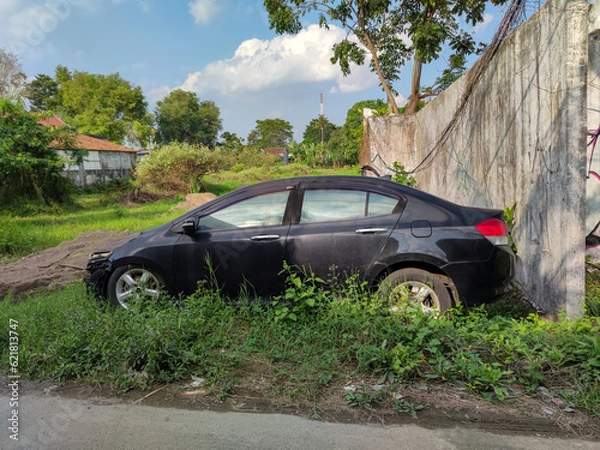 Fototapeta A black car lies abandoned in the bushes, a silent witness of a mysterious accident. The car is badly damaged, with dents, scratches and rusty. 