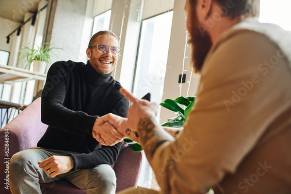 Fototapeta happy entrepreneur in eyeglasses and black turtleneck shaking hands and closing deal with tattooed businessman pointing with finger on blurred foreground, successful partnership concept