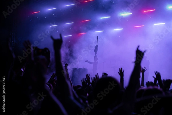 Fototapeta A group of people enjoy an illuminated music festival with raised arms. Concert Crowd in front of bright blue stage lights