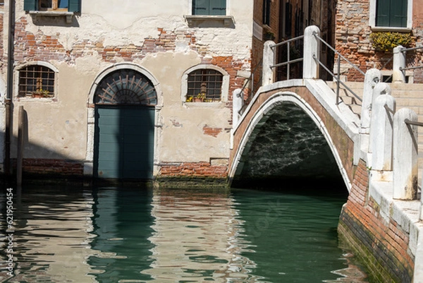 Fototapeta Shabby old walls over the water of a canal in Venice. A fragment of a building and a bridge with crumbling plaster, green shutters on windows and arched door.