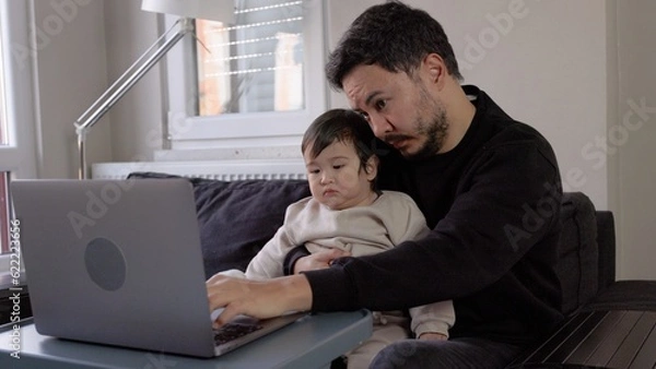 Obraz A young man trying to work on laptop holding his son at home. Remote work