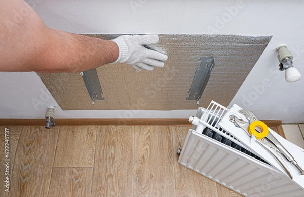 Fototapeta Foil thermal insulation on the wall behind the heating radiator at home. Thermal insulation of the apartment, to save money on utilities, heating. Energy-saving technology. 