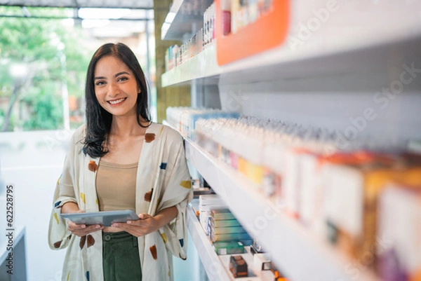 Fototapeta smiling vape seller standing beside the store shelf while holding the digital tablet