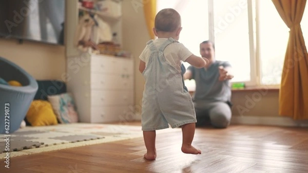 Fototapeta baby first steps. baby goes to her father at window learns to walk to take first steps. happy family kid dream concept. dad calls son baby first steps indoors. happy family lifestyle indoors concept