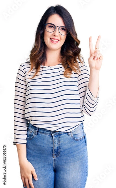Fototapeta Beautiful young brunette woman wearing casual clothes and glasses showing and pointing up with fingers number two while smiling confident and happy.