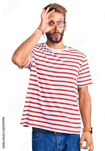 Fototapeta Handsome blond man with beard wearing casual clothes and glasses doing ok gesture shocked with surprised face, eye looking through fingers. unbelieving expression.