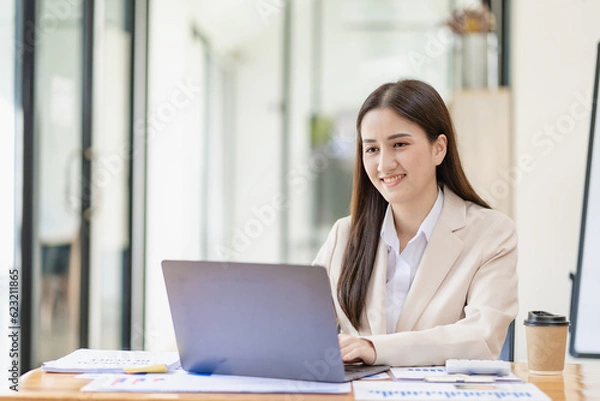 Obraz Asian female entrepreneur working in finance at home office analyzing financial graphs on documents with laptop on table in office