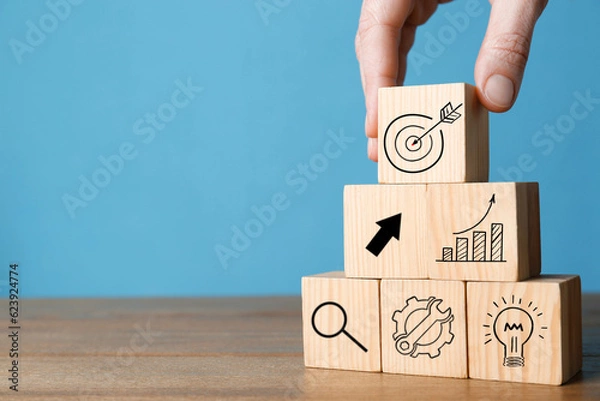 Fototapeta Woman building pyramid of cubes with icons on wooden table against light blue background, closeup. Target, arrow, magnifier and other illustrations