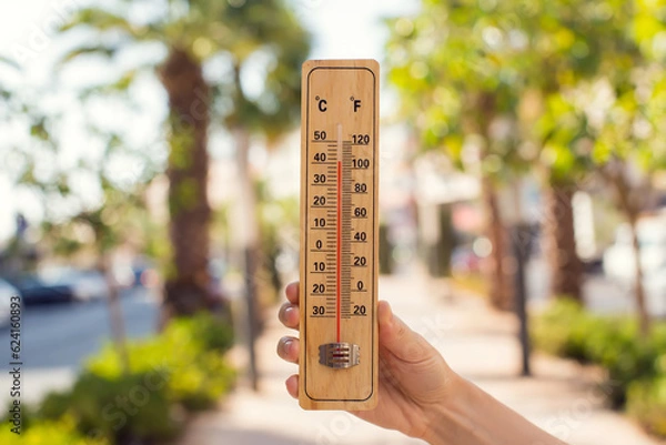 Fototapeta Hot weather. Thermometer in hand in front of street with trees and palms during heatwave. High temperature concept