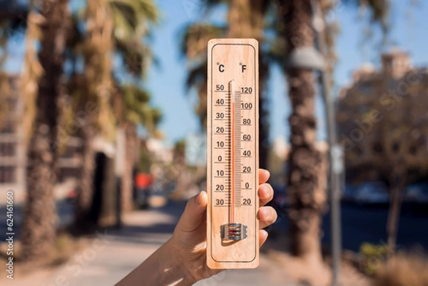 Fototapeta Hot weather. Thermometer in hand in front of street with trees and palms during heatwave. High temperature concept