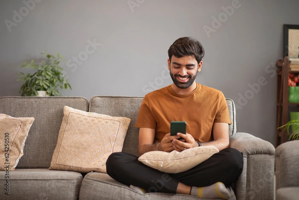 Fototapeta Young man using mobile phone while relaxing on sofa in living room