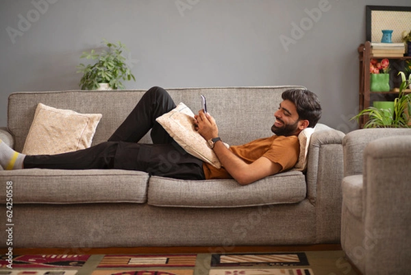 Fototapeta Young man using mobile phone while relaxing on sofa in living room