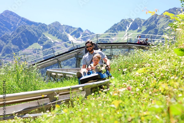 Obraz father and child having ride on summer toboggan called Rodelbahn rushing down the track . Beautiful mountains on background