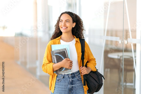 Fototapeta Woman university student. Happy lovely brazilian or hispanic female student, with a backpack, hold books and notebooks in her hand, stand near the university campus, looks and smile to the side