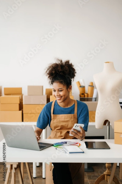 Fototapeta Startup small business SME, Entrepreneur owner African woman using smartphone or tablet taking receive and checking online purchase shopping order to preparing pack product box. in office.