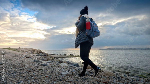 Fototapeta Female hiker in warm clothing equipped with a backpack and thermos, enjoying a walk on a rocky sea beach. A perfect addition to any travel or tourism project.