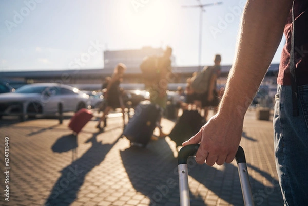 Fototapeta Group of people walking to airport terminal at summer sunset. Selective focus on hand of man with suitcase. .