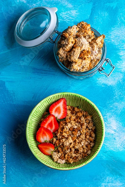 Obraz Healthy breakfast with granola and fresh strawberries served in a bowl on a blue background. Minimalistic composition with plate, spoon and natural morning light.