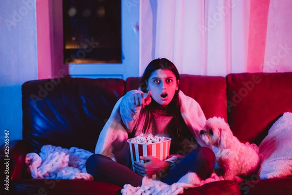 Fototapeta Beautiful young girl at home in a dark room with a popcorn bucket playing with her dog and sits in front of a monitor or TV.