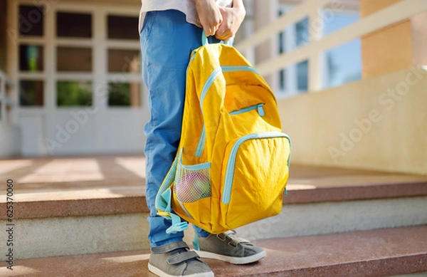 Fototapeta Little student with a backpack on the steps of the stairs of school building. Close-up of child legs, hands and schoolbag of boy standing on staircase of schoolhouse.Back to school concept.