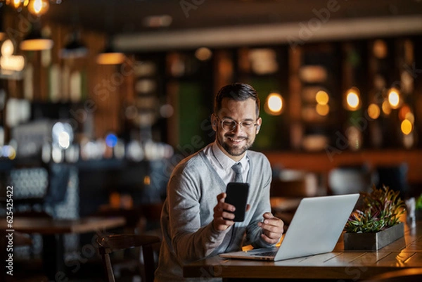 Fototapeta A happy young elegant businessman in smart casual is sitting in a coffee shop with his laptop and smiling at the phone.