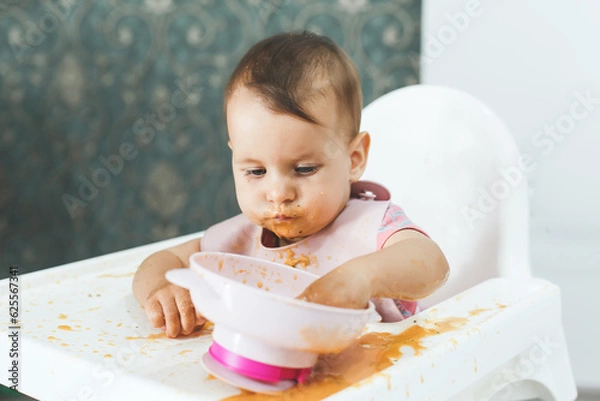 Fototapeta grimy dirty baby 9 months old sits in a highchair and eats complementary foods vegetable puree soup spilled from a plate, the concept of baby food