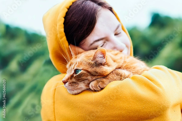 Obraz Beautiful pretty girl hugging a cute ginger cat on a blurred background. Close up portrait of a young hipster woman hugging her good friend ginger cat. Relaxation, pleasure. People and animals in love